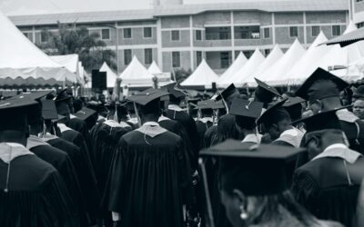 Diverse group of graduates in gowns at an outdoor ceremony with tents and buildings.